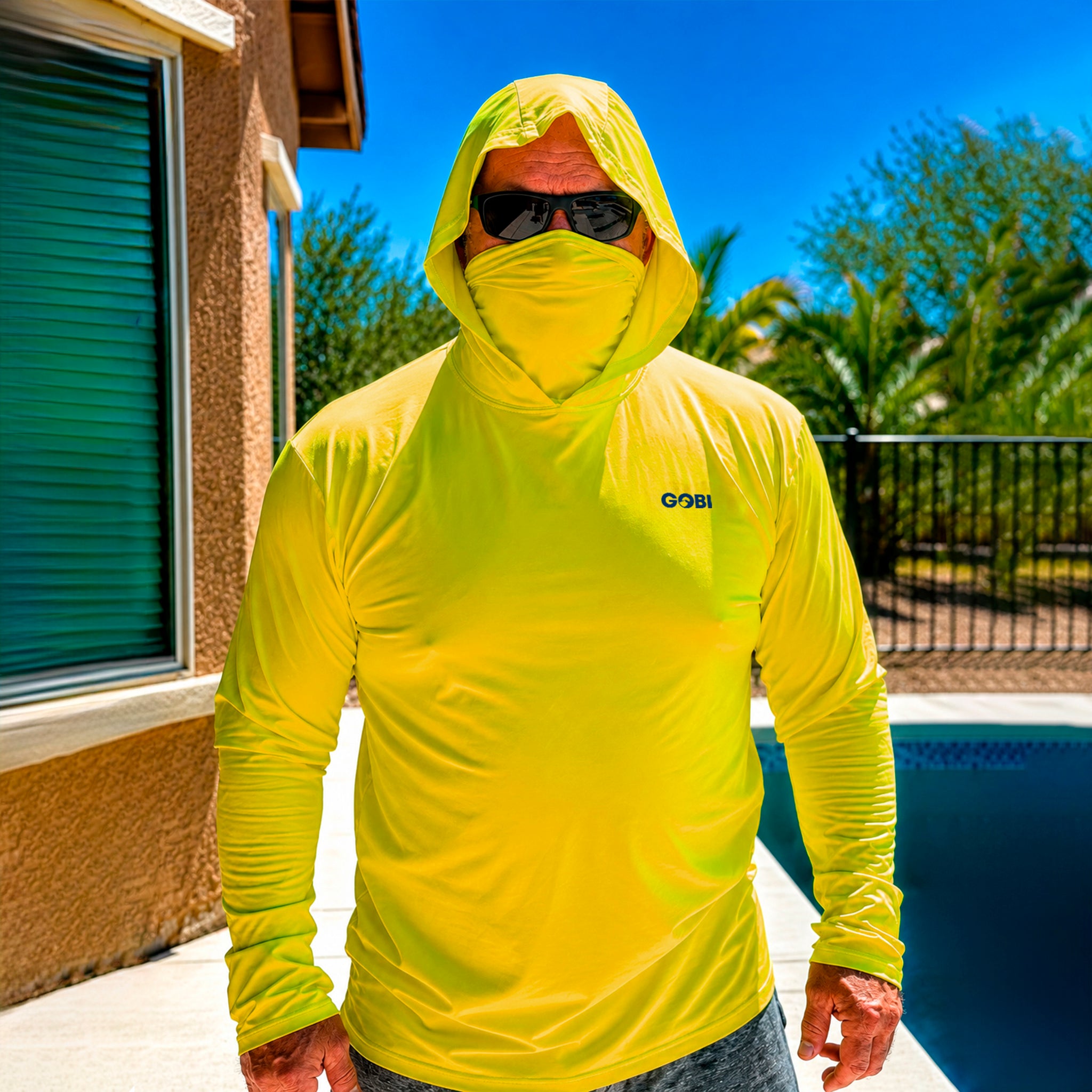 Man standing poolside in a hi-vis yellow Gobi Helio UPF Sun Hoodie with hood and face cover up, wearing sunglasses with a pool and palm trees behind him.