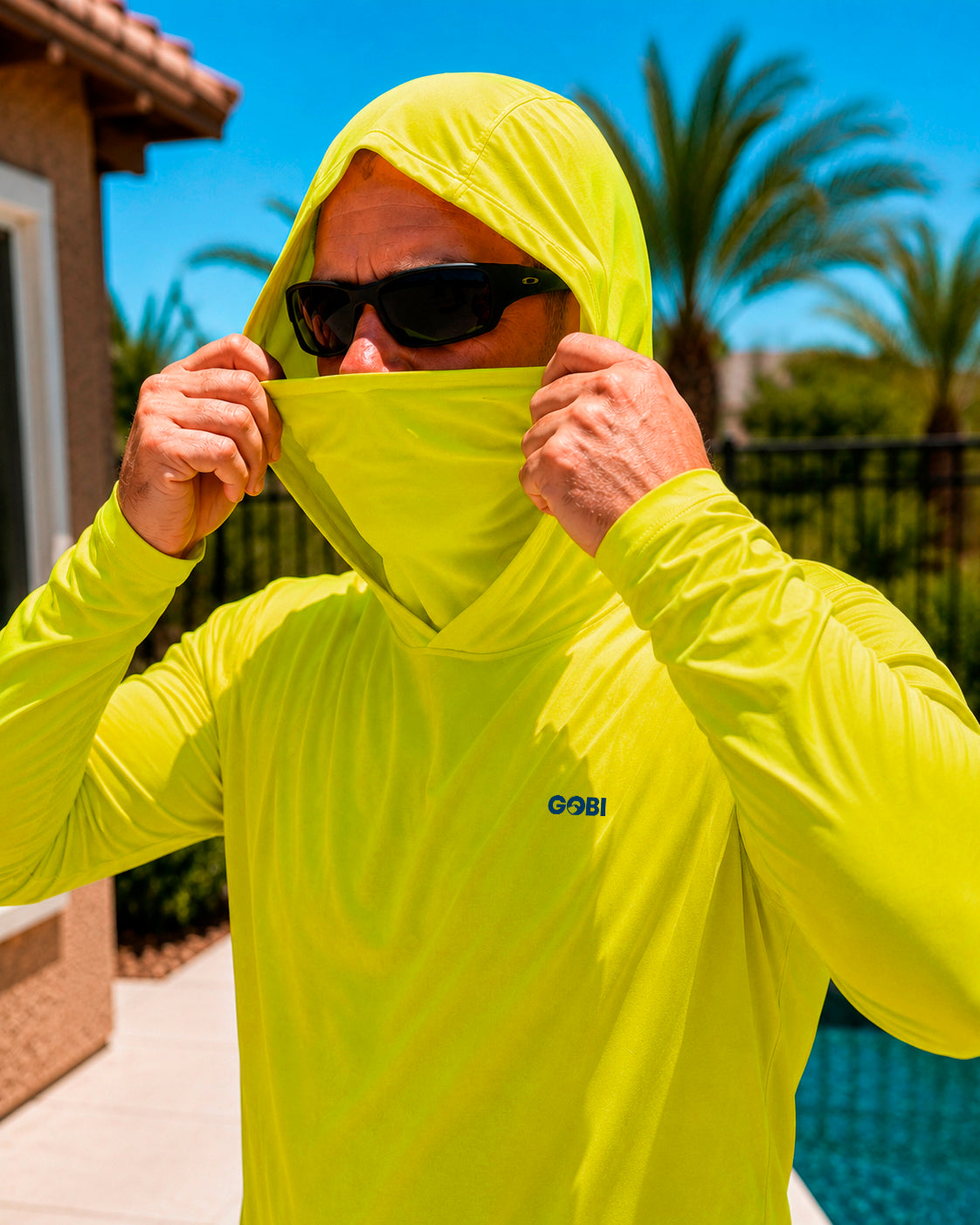 Close-up of a man pulling up the built-in face cover on a hi-vis yellow GOBI Helio UPF Sun Hoodie, wearing sunglasses outdoors with palm trees in the background.