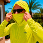 Close-up of a man pulling up the built-in face cover on a hi-vis yellow GOBI Helio UPF Sun Hoodie, wearing sunglasses outdoors with palm trees in the background.
