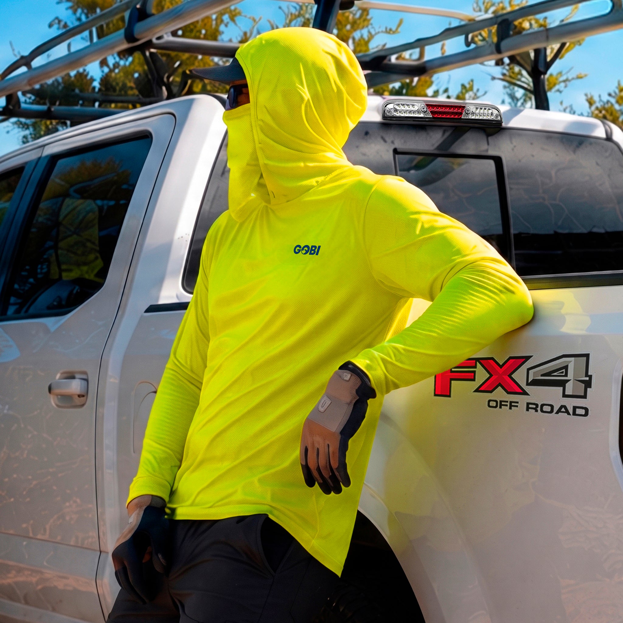 Man in a hi-vis yellow GOBI Helio UPF Sun Hoodie with hood and face cover up, leaning against a Ford FX4 Off Road pickup truck.